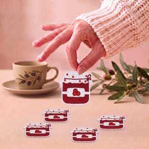 Könnte beinhalten: Ein Set aus gestickten Aufnähern in Form von Gläsern mit roter Kirschmarmelade. Jeder Aufnäher zeigt einen weißen Deckel mit roten Punkten, einen roten Glaskörper und ein weißes Etikett mit drei roten Kirschen. Detaillierte Stickerei mit rotem und weißem Garn.
