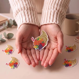 Puede incluir: Un parche de cornucopia con un diseño bordado de un cuerno dorado rebosante de una calabaza naranja, una manzana roja, uvas moradas y hojas verdes. El parche tiene un borde blanco y se muestra en varias instancias sobre una superficie clara.