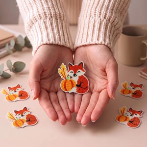 May include: An embroidered patch featuring a smiling orange and white fox sitting next to a bright orange pumpkin and a yellow wheat stalk. The patch has a white border and is rendered in a cartoon style with detailed stitching throughout.