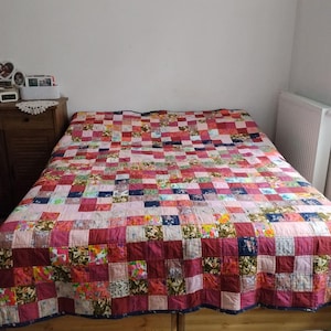 Może przedstawiać: Narzuta typu patchwork z siatką małych kwadratów w różnych odcieniach różu, czerwieni i burgunda, przeplatanych kwadratami z wielokolorowymi wzorami kwiatowymi i roślinnymi, rozłożona na drewnianej ramie łóżka.