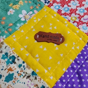 Op de afbeelding: Een patchwork quilt met diverse bloemrijke en geometrische stoffen vierkanten in turquoise, rood, geel en paars. Een bruin, leerachtig label is op het middelste gele vierkant gestikt met de tekst Hand made with Love en een hartje.