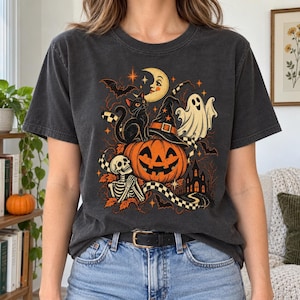 Op de afbeelding: Een antracietgrijs t-shirt met een grafische print van een pompoen, een zwarte kat, een skelet, een geest, een maansikkel, vleermuizen en een spookhuis. Het ontwerp bevat oranje, witte en zwarte kleuren met een geblokt patroon.