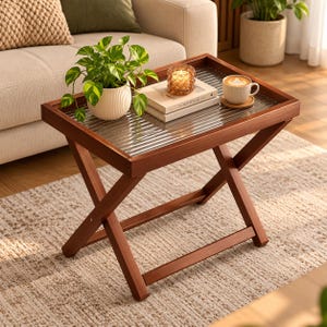 May include: A wooden folding tray table with a rectangular frame and a ribbed glass top. On the table sits a potted green plant in a white ribbed ceramic pot, a stack of two books, a glass candle holder, and a white ceramic coffee mug.