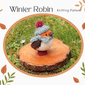 Könnte beinhalten: Strickanleitung für einen Winter-Rotkehlchen, einen kleinen gestrickten Vogel, der einen grauen Hut mit rotem Bommel trägt. Das Rotkehlchen sitzt auf einer Holzscheibe.
