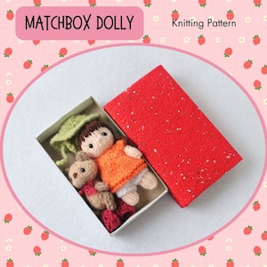 May include: A knitted doll and a teddy bear are inside a matchbox. The doll is wearing a green hat, an orange sweater, and white pants. The teddy bear is brown with a red bow. The matchbox is red with white speckles. The image is on a pink background with white flowers and red strawberries.