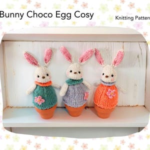 May include: Three knitted bunny egg cozies in a row. Each bunny has a white body, pink-lined ears, and a sweater in teal, lavender, and orange. The bunnies sit in terracotta pots. The text "Bunny Choco Egg Cosy" and "Knitting Pattern" are at the top.