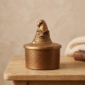 以下が含まれることがあります： 金属製のゴールドカラーの円筒形容器で、蓋には彫刻が施された使い古した帽子のようなデザインがあしらわれています。容器の側面にはレンガ模様のテクスチャがエンボス加工されており、細部まで作り込まれた均一な外観に仕上がっています。