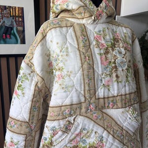 May include: A quilted hooded sweatshirt featuring a floral pattern with pink, blue, and green flowers on a white background with tan border accents. The garment has a large front kangaroo pocket and a hood with a gathered, elasticised neckline.