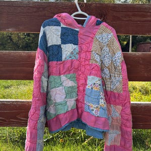 May include: A hooded long-sleeve pullover constructed from a patchwork quilt featuring various fabric squares in shades of pink, blue, green, and tan. The garment has a textured, quilted surface with visible stitching throughout the fabric panels.