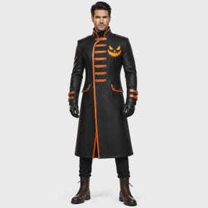Op de afbeelding: Een zwarte lange jas met oranje biezen, een hoge kraag en een afbeelding van een pompoengezicht op de borst. De jas heeft horizontale oranje banden aan de voorzijde, bijpassende oranje manchetten, zwarte handschoenen, een zwarte broek en bruine laarzen.