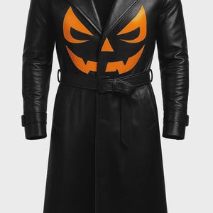 Könnte beinhalten: Ein schwarzer Trenchcoat mit einem großen, orangefarbenen Kürbisgesicht-Motiv auf der Brust. Der Mantel verfügt über einen breiten Kragen, lange Ärmel mit Schnallen an den Manschetten und einen passenden schwarzen Gürtel mit Schnalle in der Taille.