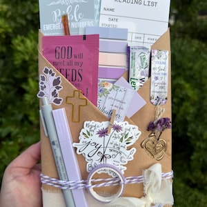 May include: A brown paper envelope filled with stationery items including a reading list card, a pink packet labeled God will meet all my needs, a cross-shaped paperclip, a purple pen, a floral bookmark, a sticker with the text the joy of the Lord is my strength, and a roll of washi tape tied with white and purple twine.