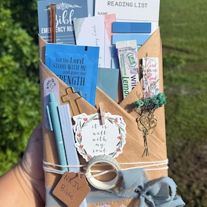 May include: A brown paper envelope containing a Bible, a reading list, blue sticky notes, a True Lemon drink mix packet, a gold cross, two light blue pens, a floral sticker, a small dried flower bouquet, a roll of washi tape, and a tag labeled NKJV Bible.