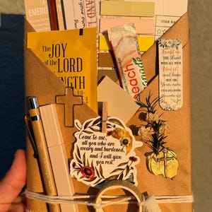 May include: A brown paper envelope containing a reading list, a yellow packet labeled The Joy of the Lord is My Strength, a peach drink mix packet, a cross-shaped paperclip, a floral bookmark, a pen, a roll of tape, and a wax seal with a cross.
