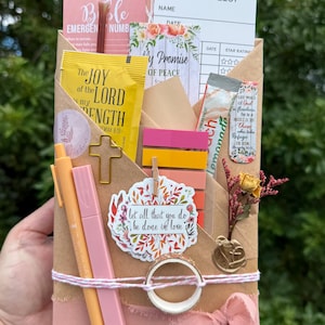 May include: A brown paper envelope filled with stationery items including a reading list card, a yellow packet labeled The Joy of the Lord is my Strength, a gold cross paperclip, colorful sticky tabs, a floral sticker, and a pink ribbon.