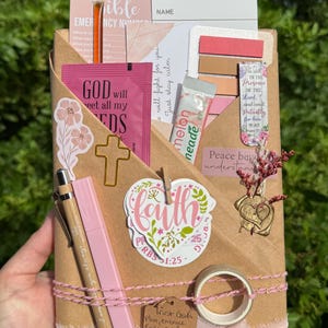 May include: A brown paper-wrapped package containing a reading list card, a pink packet labeled God will meet all my needs, a melon drink mix, a cross-shaped paperclip, a faith sticker, a pen, a highlighter, sticky notes, and a wax seal.