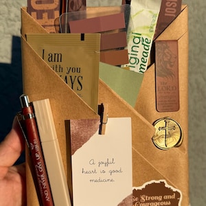 May include: A brown paper envelope containing various items including a red pen with text, a beige pen, a card reading A joyful heart is good medicine, a gold wax seal with a cross, and several bookmarks with religious text and lion imagery.
