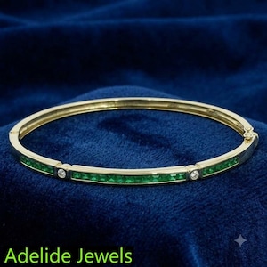 May include: A gold hinged bangle bracelet featuring a continuous row of square-cut green emeralds accented by two round brilliant-cut diamonds set in gold. The piece includes a secure clasp mechanism and the text Adelide Jewels is visible.