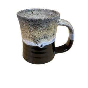 Peut inclure: Une tasse en céramique avec un glaçage noir, blanc et brun. La tasse a un corps arrondi et une anse.