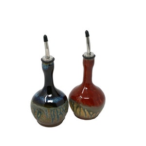 May include: Two ceramic oil and vinegar bottles with metal pour spouts. The bottle on the left is blue and green with a brown base. The bottle on the right is red with a brown base.