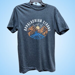 Puede incluir: Una camiseta gris oscuro con un gr&aacute;fico de monta&ntilde;as y un sol poni&eacute;ndose detr&aacute;s de ellas. El texto "Appalachian Strong" est&aacute; escrito alrededor del gr&aacute;fico.