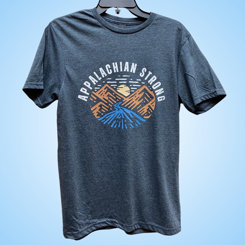 Puede incluir: Una camiseta gris oscuro con un gr&aacute;fico de monta&ntilde;as y un sol poni&eacute;ndose detr&aacute;s de ellas. El texto "Appalachian Strong" est&aacute; escrito alrededor del gr&aacute;fico.