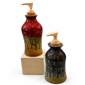 Könnte beinhalten: Zwei Keramik-Seifenspender mit tropfendem Glasurdesign. Einer ist rot, grün und gelb, der andere dunkelblau und grün. Beide haben einen Korkverschluss und eine beige Pumpe.