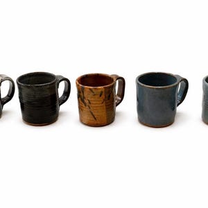 May include: Five handcrafted ceramic mugs in different colours and textures. The mugs include a black and grey mug, a dark grey mug, a brown mug, a blue mug, and a grey mug. Each mug has a handle and a unique design.