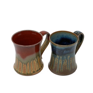 May include: Two ceramic mugs with drip glaze designs. One mug is red with a yellow and green drip glaze. The other mug is blue with a brown and yellow drip glaze.