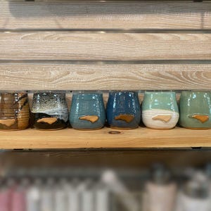 May include: A collection of six ceramic stemless wine glasses in various colors, including brown, black, teal, and green. Each glass features a brown, North Carolina state-shaped accent. The glasses are displayed on a wooden shelf against a wooden background.
