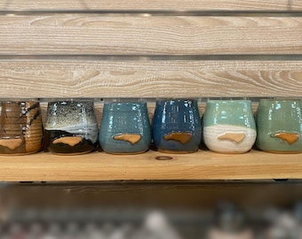 May include: Six ceramic stemless wine glasses in shades of brown, black, teal, and green. Each glass has a brown, North Carolina state-shaped detail. The glasses are displayed on a wooden shelf against a wooden backdrop. The glasses are suitable for approximately 350 ml.