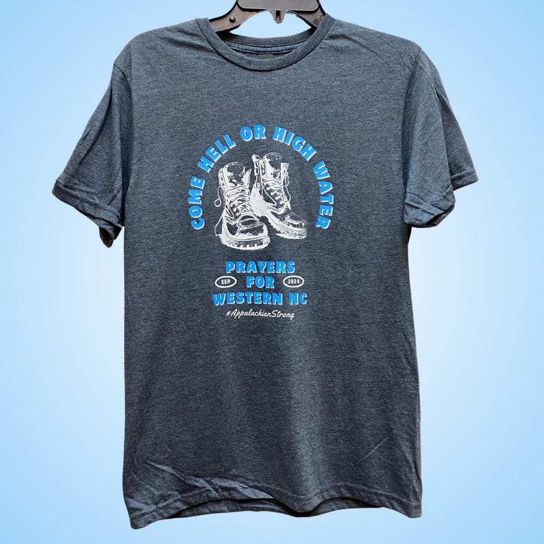 Puede incluir: Una camiseta gris oscuro con un gr&aacute;fico blanco de dos botas de senderismo y el texto "Come Hell or High Water, Prayers for Western NC, #AppalachianStrong".