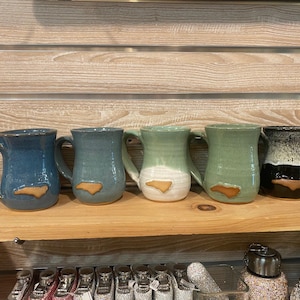 May include: A collection of ceramic mugs in various colors, including blue, green, and black, each featuring a tan-colored outline of a state. The mugs are displayed on a wooden shelf.