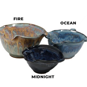 May include: Three ceramic mixing bowls in different sizes and colours. The largest bowl is a light blue with brown and orange streaks and is labelled "FIRE". The medium bowl is a light blue with darker blue streaks and is labelled "OCEAN". The smallest bowl is a dark blue and is labelled "MIDNIGHT".