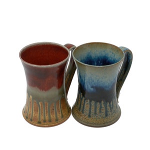 May include: Two ceramic mugs with drip glaze designs. One mug is red and brown with a light brown drip glaze. The other mug is blue and gray with a light brown drip glaze.