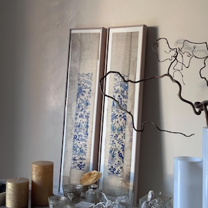 Peut inclure: Deux cadres muraux rectangulaires et hauts présentant des illustrations verticales bleues et blanches de personnages et de paysages sur un fond beige texturé, exposés avec des bougies piliers dorées et des récipients en verre.