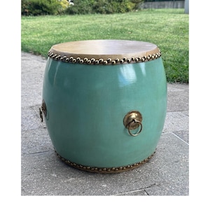 May include: A barrel-shaped stool with a smooth, light green finish. The top and bottom edges are decorated with a row of small, metallic brass-colored studs. Two circular brass ring handles are mounted on the sides of the cylindrical body.