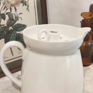 Può includere: Una brocca in ceramica bianca con un corpo largo e arrotondato, un manico curvo sul lato e un coperchio aderente dotato di una piccola impugnatura circolare. La brocca presenta una finitura liscia e lucida e un beccuccio distinto.