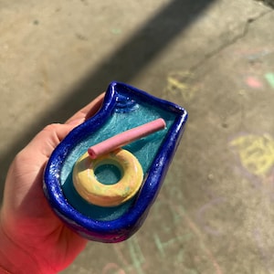 May include: A small, handmade clay sculpture of a rectangular swimming pool with a glossy dark blue border and a light blue interior. Inside the pool, there is a yellow circular pool float and a small, pink cylindrical object resting on the surface.