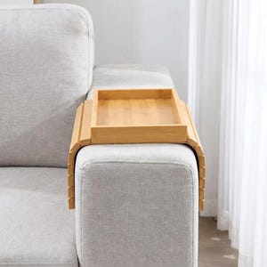 May include: A light brown bamboo sofa arm tray featuring a flat rectangular surface and flexible, slatted sides that drape over the arm of a couch. The tray is constructed from natural wood segments, providing a stable platform for items.