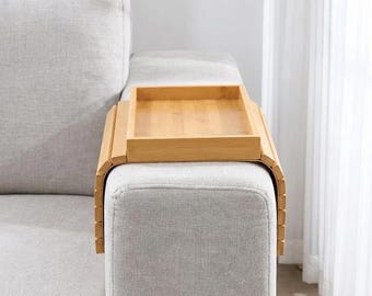 May include: A light brown bamboo sofa arm tray featuring a flat rectangular surface and flexible, slatted sides that drape over the arm of a couch. The tray is constructed from natural wood segments, providing a stable platform for items.