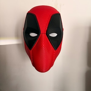 Puede incluir: Una máscara facial completa de color rojo y negro con un patrón texturizado similar a la fibra de carbono en la superficie roja. La máscara incluye grandes parches oculares negros en forma de almendra con lentes de malla blanca.