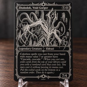 May include: A black and white trading card featuring the character Zhulodok, Void Gorger. The card displays a detailed illustration of a multi-limbed creature, the text 'Legendary Creature — Eldrazi', and game rules regarding colourless spells.