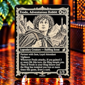 May include: A black and white collectible trading card titled Frodo, Adventurous Hobbit. The card features an illustration of a curly-haired character in a cloak, a mountain landscape, and game text including Legendary Creature, Halfling Scout.
