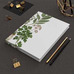 May include: A white hardback journal featuring a botanical illustration of green herbs and brown seed heads on the front cover. The notebook is positioned alongside two pencils, one gold and one black, and several gold metal binder clips.