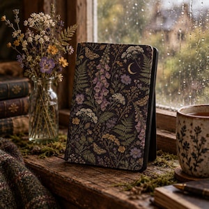 May include: A black e-reader case featuring a detailed botanical print of purple foxgloves, ferns, wildflowers, moths, and a golden crescent moon. The case stands upright on a wooden surface next to a glass vase of dried flowers and a ceramic mug.