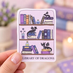 May include: A rectangular sticker with a light purple background featuring three shelves of books, small potion bottles, and three black dragons. The text "LIBRARY OF DRAGONS" is printed in dark purple at the bottom of the sticker design.