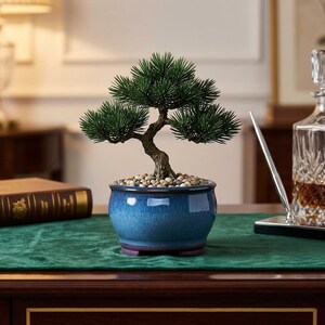 May include: A small artificial bonsai tree with dark green needle-like foliage and a textured brown trunk. The tree is planted in a round, glossy ceramic pot with a blue gradient finish, filled with small, light-colored decorative pebbles.