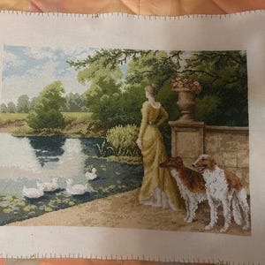 May include: A rectangular cross-stitch embroidery on white fabric featuring a landscape scene with a person in a yellow dress standing by a stone wall, two brown and white dogs, a pond with white ducks, green trees, and a stone planter.