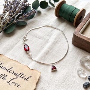 Op de afbeelding: Een zilveren kettingarmband met een marquise-geslepen rode edelsteen en een kleinere driehoekige rode edelsteenbedel. De armband heeft een ronde sluiting en wordt getoond naast een houten doosje en takjes gedroogde lavendel.